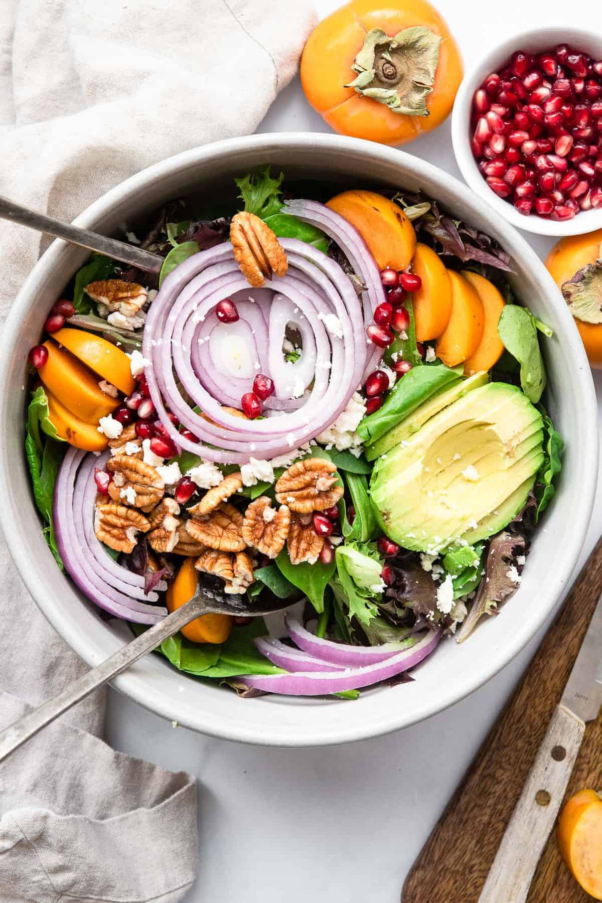 Pomegranate Persimmon Salad - This fall salad is bright and colorful with persimmons, feta and the yummiest honey balsamic vinaigrette. A quick and easy, healthy salad that everyone will want the recipe for! | #Foodfaithfitness | #glutenfree #highprotein #grainfree #healthy #salad pomegranate persimmon salad in a ceramic bowl with salad tongs