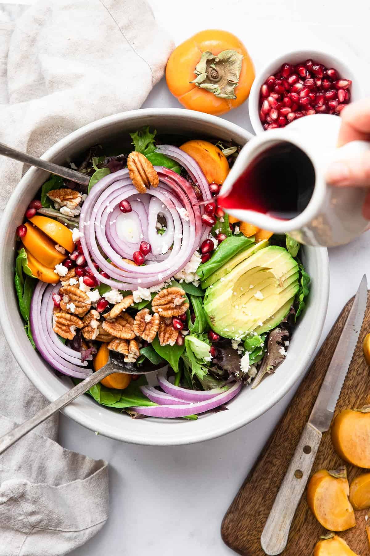 Pomegranate Persimmon Salad - This fall salad is bright and colorful with persimmons, feta and the yummiest honey balsamic vinaigrette. A quick and easy, healthy salad that everyone will want the recipe for! | #Foodfaithfitness | #glutenfree #highprotein #grainfree #healthy #salad a hand pouring dressing over a bowl of pomegranate persimmon salad
