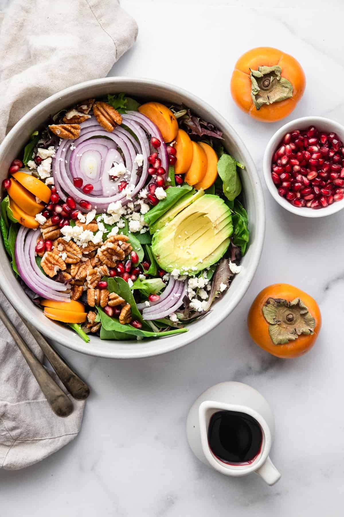 Pomegranate Persimmon Salad - This fall salad is bright and colorful with persimmons, feta and the yummiest honey balsamic vinaigrette. A quick and easy, healthy salad that everyone will want the recipe for! | #Foodfaithfitness | #glutenfree #highprotein #grainfree #healthy #salad persimmon fruit salad in a gray bowl with dressing on the side