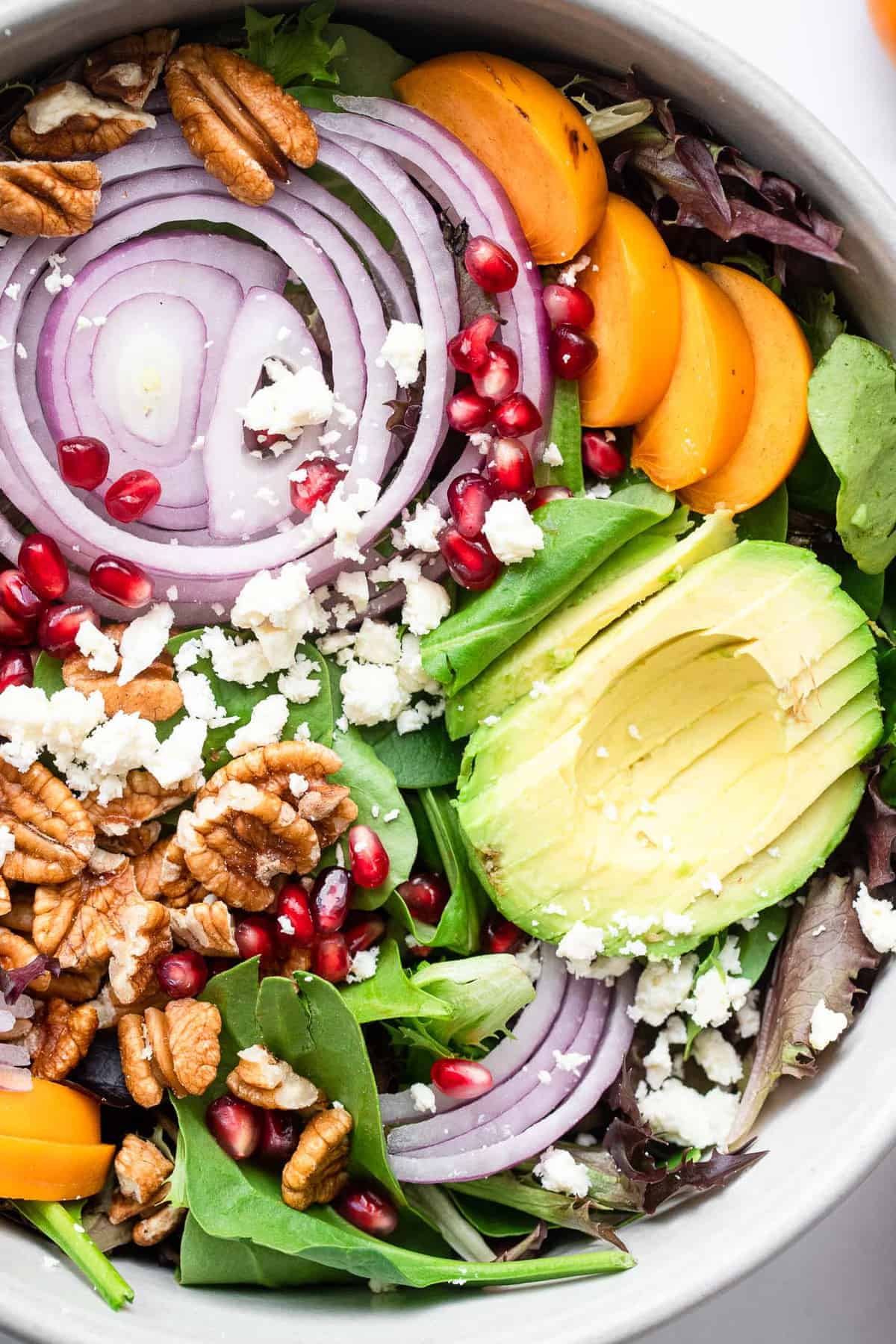 Pomegranate Persimmon Salad - This fall salad is bright and colorful with persimmons, feta and the yummiest honey balsamic vinaigrette. A quick and easy, healthy salad that everyone will want the recipe for! | #Foodfaithfitness | #glutenfree #highprotein #grainfree #healthy #salad close up of persimmon pomegranate salad in a gray bowl