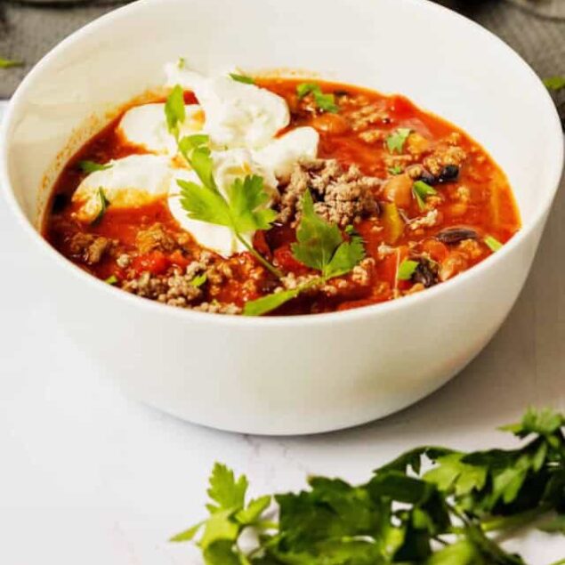 Hearty homemade chili with ground meat, tomatoes, and beans garnished with fresh cilantro and a dollop of sour cream in a white bowl.