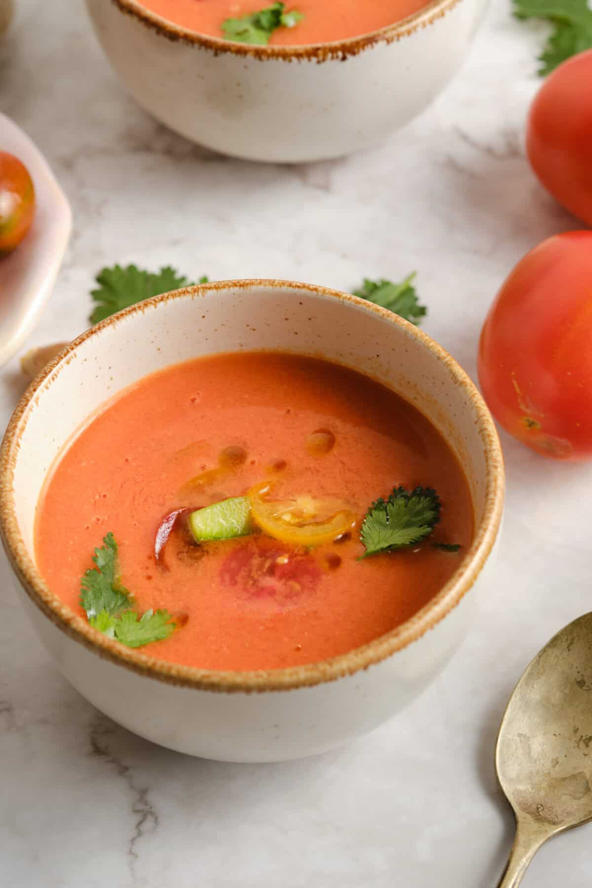 A bowl of gazpacho, garnished with fresh cilantro, sliced tomatoes, and olive oil.
