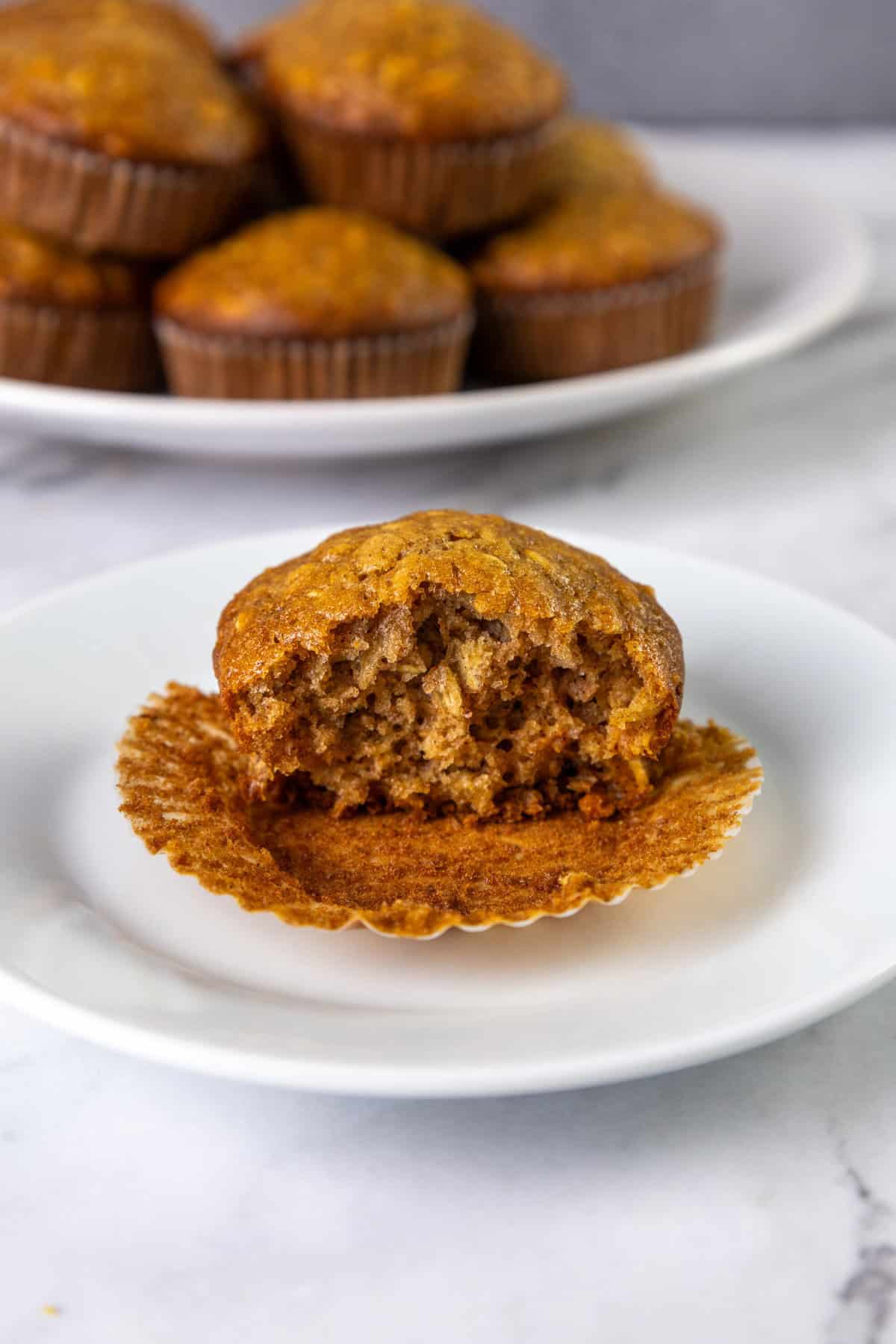 A partially eaten oatmeal muffin on a white plate, with more muffins in the background.