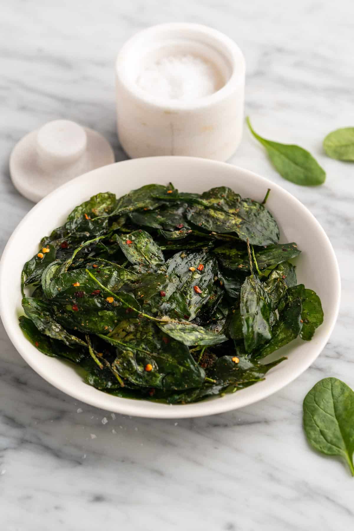 A white bowl filled with crispy spinach chips, seasoned with red pepper flakes, on a marble surface.