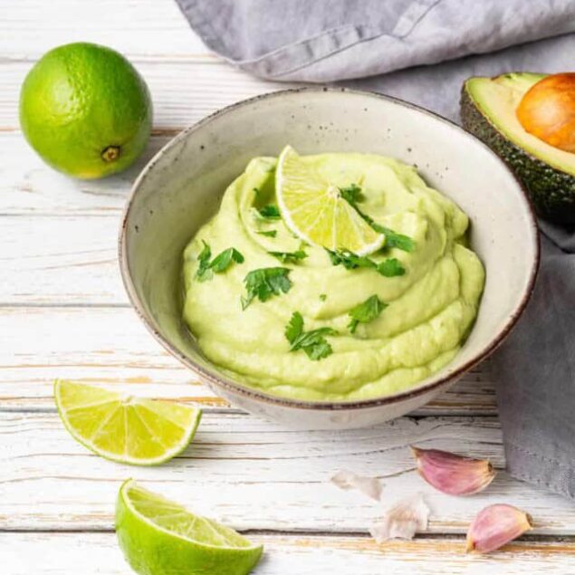 Creamy avocado guacamole with fresh lime slices and herbs in a rustic bowl on a white wooden table, featuring ingredients like lime and garlic for healthy eating and delicious snacks.