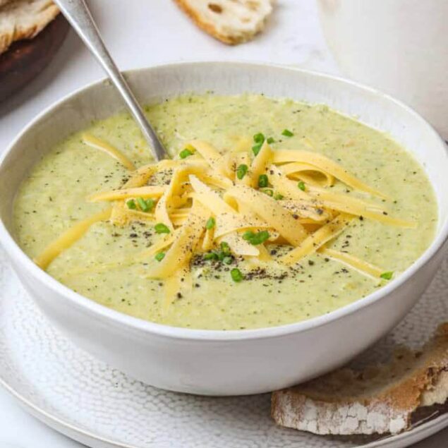 Creamy zucchini soup with shredded cheese and chopped green onions, served in a white bowl with crusty bread on the side, perfect for a healthy, comforting meal.