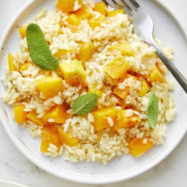 Creamy rice risotto with butternut squash and fresh sage leaves on a white plate, topped with black pepper, featuring healthy grain and vegetable ingredients for a nourishing meal.