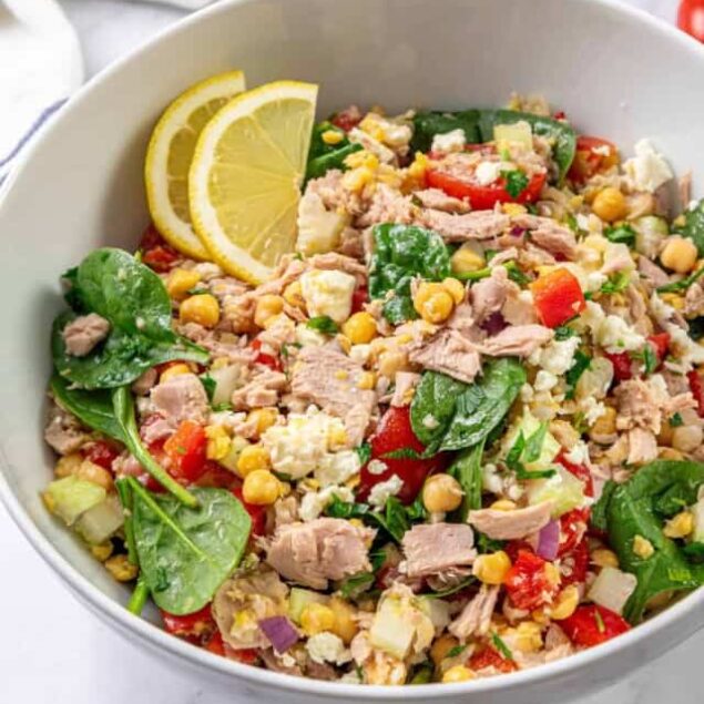 Fresh tuna salad with leafy spinach, cherry tomatoes, corn, red onion, and crumbled cheese served with lemon slices in a white bowl. Perfect for a healthy, high-protein meal or lunch.