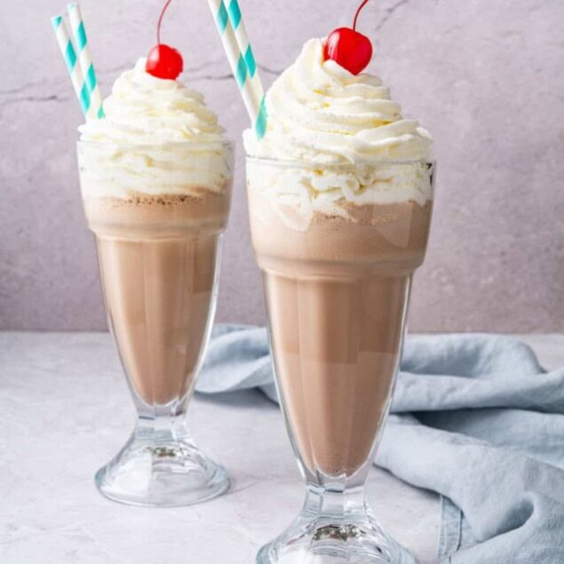 Creamy chocolate milkshake with whipped cream, maraschino cherry, and striped straws in tall glasses, perfect for dessert or a snack.