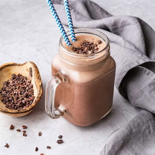 Creamy chocolate smoothie in a glass jar with blue and white striped straws, served on a light gray surface with cacao nibs and a gray cloth, promoting healthy, delicious recipes.