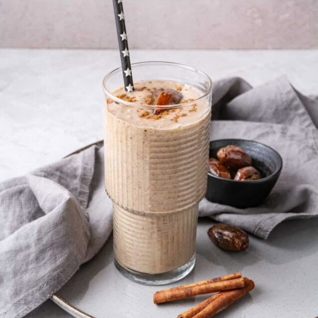 Creamy date and cinnamon plant-based protein smoothie in a glass with a black star-patterned straw, served on a ceramic plate with cinnamon sticks and Medjool dates, perfect for healthy living and fitness.