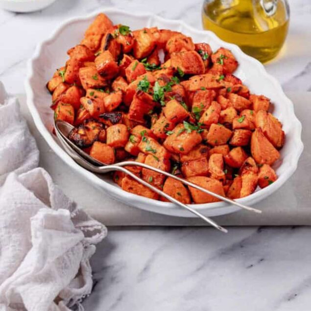Crispy roasted sweet potato cubes garnished with fresh herbs in a white bowl, with olive oil and chopped green onions on the side, perfect for healthy meal recipes.