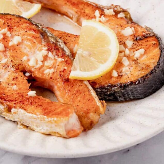 Grilled salmon fillets with lemon wedges, garnished with chopped garlic on a white plate, healthy seafood dish, high-protein meal, perfect for nutritious eating.