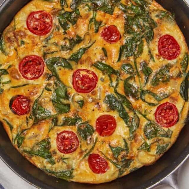 Golden baked spinach and cherry tomato frittata in a black skillet, healthy breakfast option with cheese, eggs, and fresh greens. Perfect for breakfast or brunch.