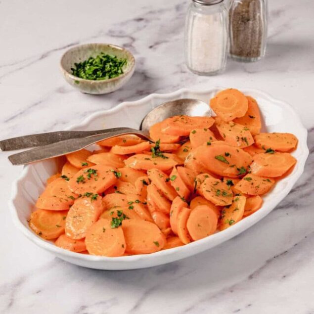 Sliced cooked sweet potatoes garnished with chopped parsley in a white oval dish on marble surface with salt and pepper shakers in the background.