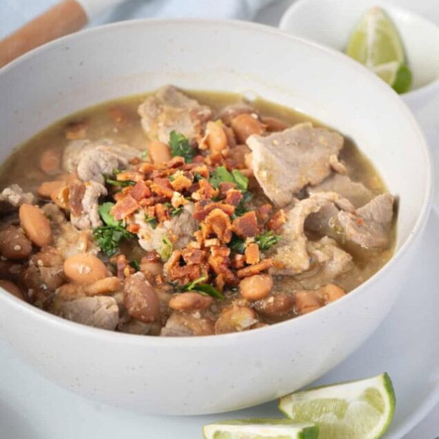 Hearty pork and bean soup with fresh herbs and crispy bacon garnish, served with lime wedges for added flavor. Perfect for a nutritious, filling meal that's easy to make at home.