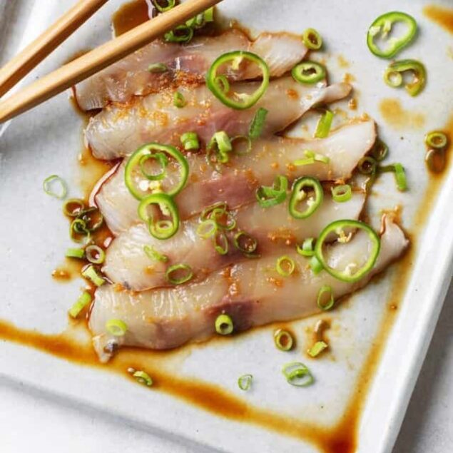 Tender raw fish slices garnished with chopped green onions on a white rectangular plate, drizzled with a savory soy-based dipping sauce, perfect for healthy, flavorful Asian-inspired cuisine.