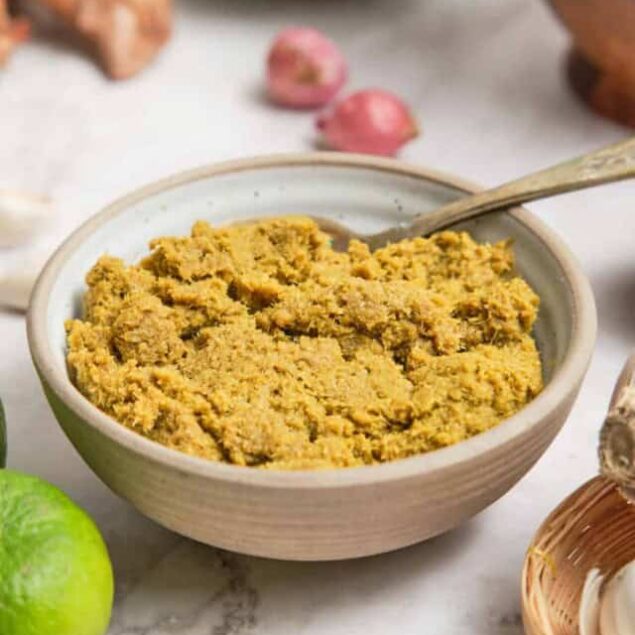 Creamy turmeric hummus in a white bowl on marble surface, garnished with herbs, surrounded by fresh ingredients like lime, garlic, and shallots for healthy eating and plant-based recipes.