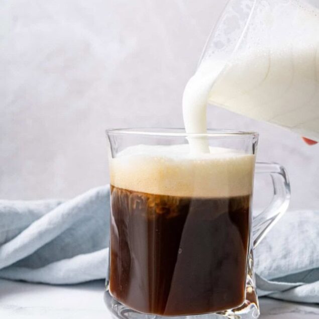 Cream being poured into a glass mug of freshly brewed black coffee.