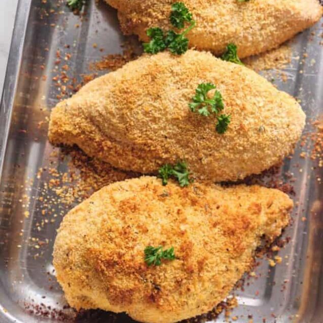 Golden baked chicken breasts with crispy breadcrumb coating garnished with fresh parsley on a baking sheet.