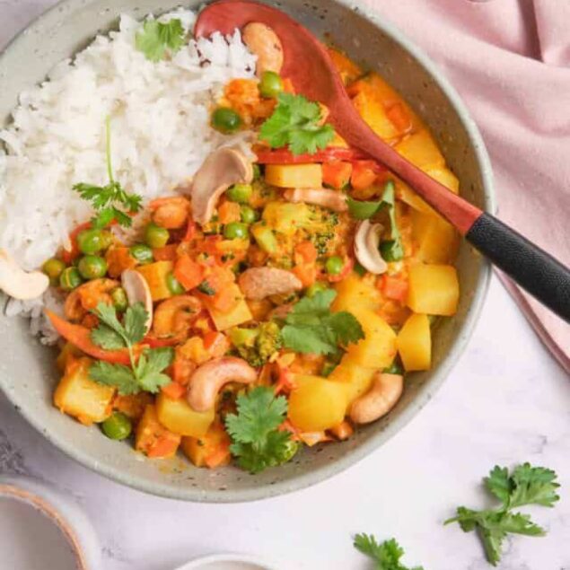 Vegan vegetable curry with rice served in a bowl, garnished with cilantro, carrots, peas, and potatoes, on a white marble surface.