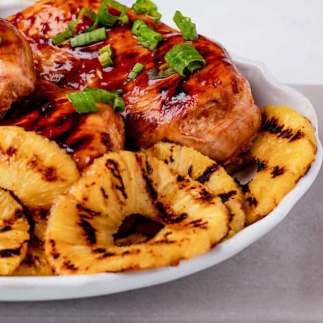 Juicy grilled chicken breast topped with barbecue sauce and garnished with chopped green onions, served with grilled pineapple rings on a white plate, perfect for healthy eating.