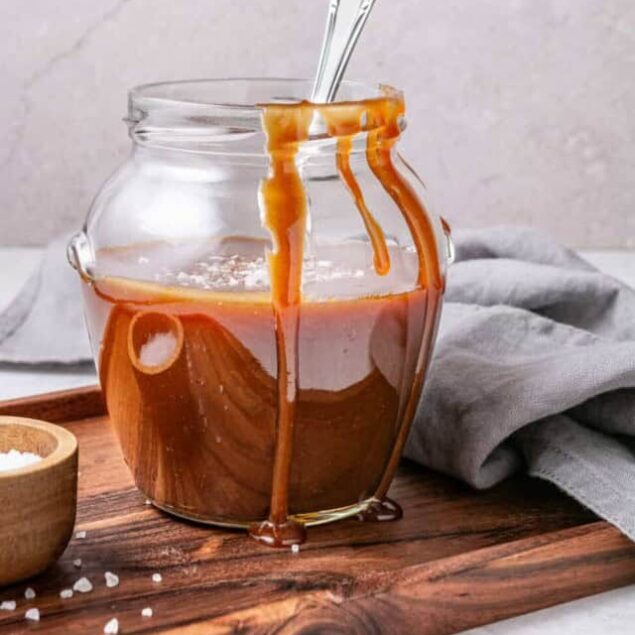 Creamy caramel sauce drizzling from a spoon into a glass jar on a wooden board, with salt and a grey cloth nearby. Perfect for drizzle recipes and sweet treats.