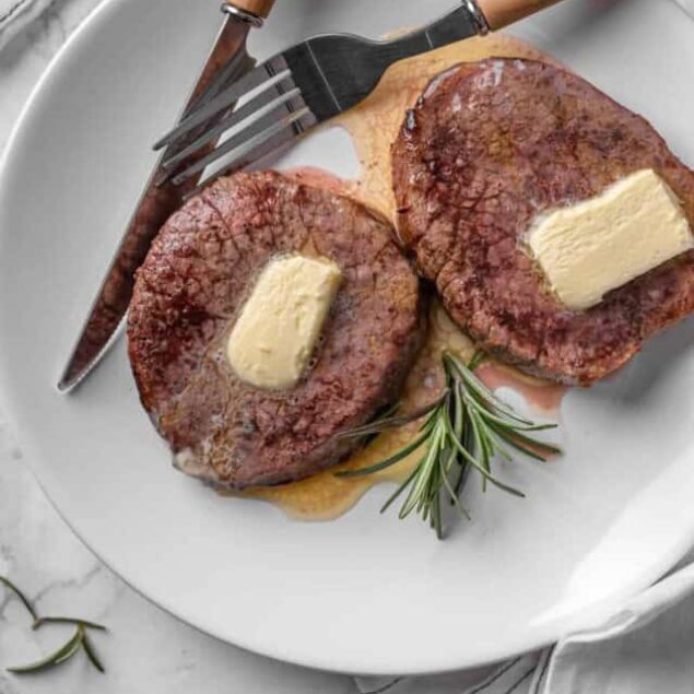 Juicy beef steaks topped with butter on a white plate with fresh rosemary, fork, and knife; perfect for healthy, high-protein meal ideas and keto-friendly dinner recipes.