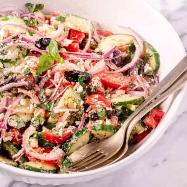 Fresh vegetable Greek salad with cucumbers, tomatoes, red onions, olives, and feta cheese in a white bowl, served with a fork, perfect for healthy eating and meal prep.