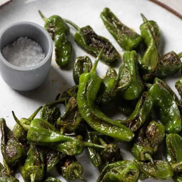 Charred green shishito peppers with salt and a dipping sauce on a white plate, showcasing a healthy, flavorful appetizer or snack option.