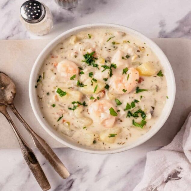 Creamy shrimp and potato chowder in a white bowl with fresh herbs, served on a marble surface with vintage utensils and seasoning shakers, perfect for cozy, wholesome meals.