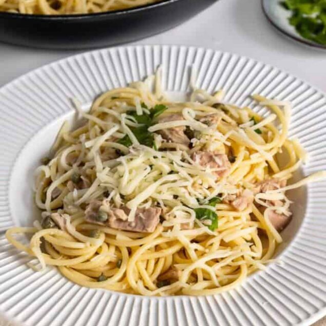 Spaghetti with tuna, cheese, and herbs on a white textured plate, close-up shot.