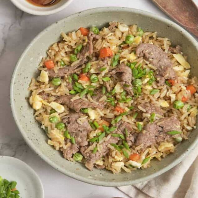 Savory beef and vegetable fried rice in a ceramic bowl with soy sauce and fresh herbs.