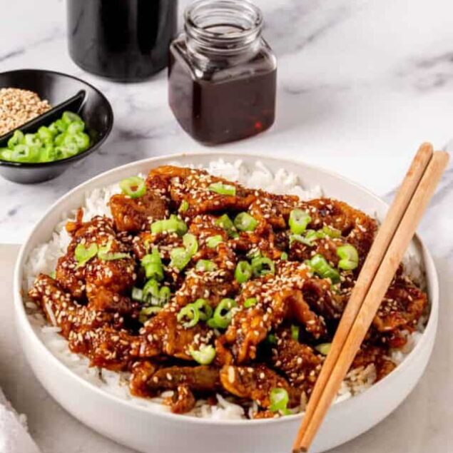 Sticky Asian chicken stir-fry with sesame seeds and green onions on a bed of white rice, served with soy sauce and garnished with chopped scallions for a healthy and delicious meal.