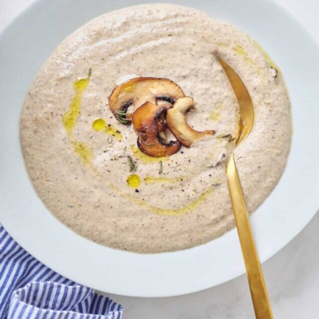 Creamy mushroom soup garnished with sautéed mushrooms and a drizzle of olive oil, served in a white bowl with a gold spoon, perfect for healthy eating and comfort food recipes.