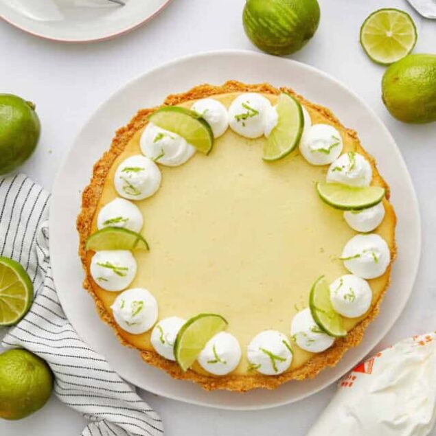 Lemon lime pie with whipped cream and lime slices on a white cake stand, surrounded by fresh limes and baking ingredients.