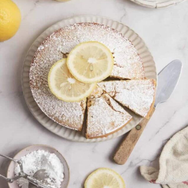 Lemon cake topped with powdered sugar and lemon slices on a white plate. Fresh lemons and eggs on a marble surface, with a cake server for serving. Perfect for lemon dessert recipes and healthy, flavorful treats.