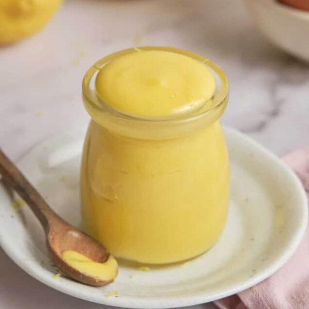 Creamy lemon curd in a glass jar on a white plate with a wooden spoon, fresh lemons in the background, bright and refreshing dessert or spread, healthy treat recipe Featured on Food Faith Fitness.
