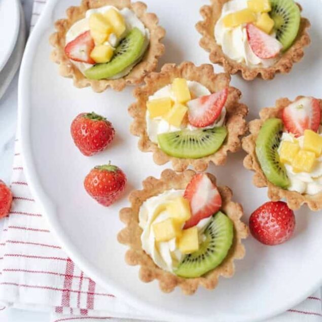 Fresh fruit tartlets with strawberries, kiwi, mango, and whipped cream on a white serving platter. Perfect healthy dessert option from Food Faith Fitness.