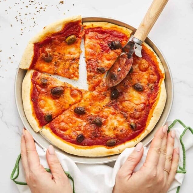 Cheese and black olive pizza on a round metal tray, being sliced with a pizza cutter, over a white marble surface, with a person's hands holding a cloth.