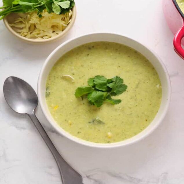 Creamy green vegetable soup with fresh cilantro garnish, served in a white bowl on a white marble surface, with a side of shredded cheese and herbs, perfect for healthy eating and meal ideas.
