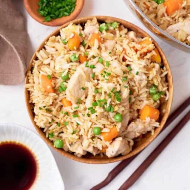 Fresh homemade chicken fried rice with vegetables, served in a wooden bowl, garnished with chopped green onions, accompanied by soy sauce and chopsticks for a healthy and flavorful meal.