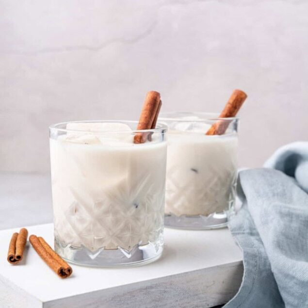Creamy vanilla milkshake with cinnamon sticks on a white table, served in textured glass cups. Perfect for healthy dessert recipes or indulgent treats on Food Faith Fitness.