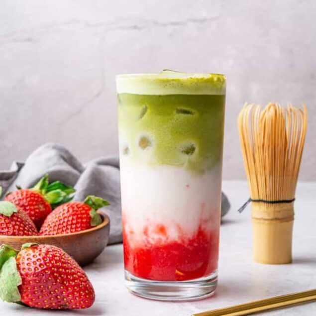 Fresh strawberry and matcha layered smoothie in a tall glass with strawberries and a bamboo whisk, healthy and delicious recipe from Food Faith Fitness.