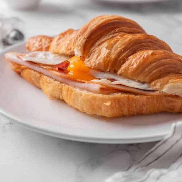 Golden croissant sandwich with egg, ham, and melted cheese on a white plate, perfect for breakfast or brunch. Fresh, buttery pastry paired with savory fillings for a delicious meal.