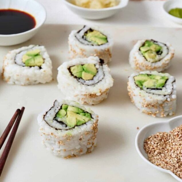 Sushi rolls with avocado and sesame seeds on a white serving board, accompanied by soy sauce, wasabi, ginger, and sesame seeds. Healthy, delicious sushi recipes for a balanced diet from Food Faith Fitness.
