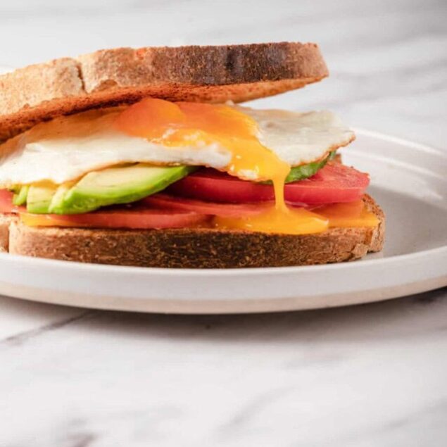 Savory breakfast sandwich with fried egg, avocado, tomato, and whole grain bread on a white plate.