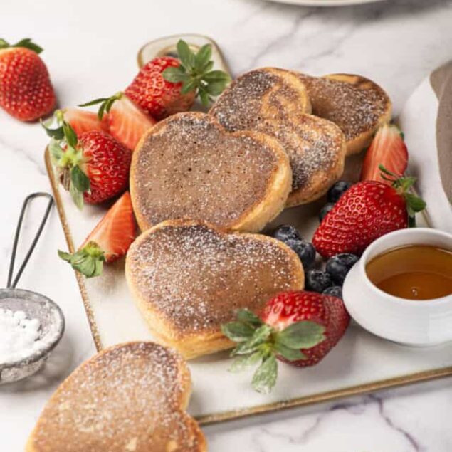 Fluffy heart-shaped cinnamon pancakes served with fresh strawberries, blueberries, and syrup on a white marble table, perfect for breakfast or brunch.