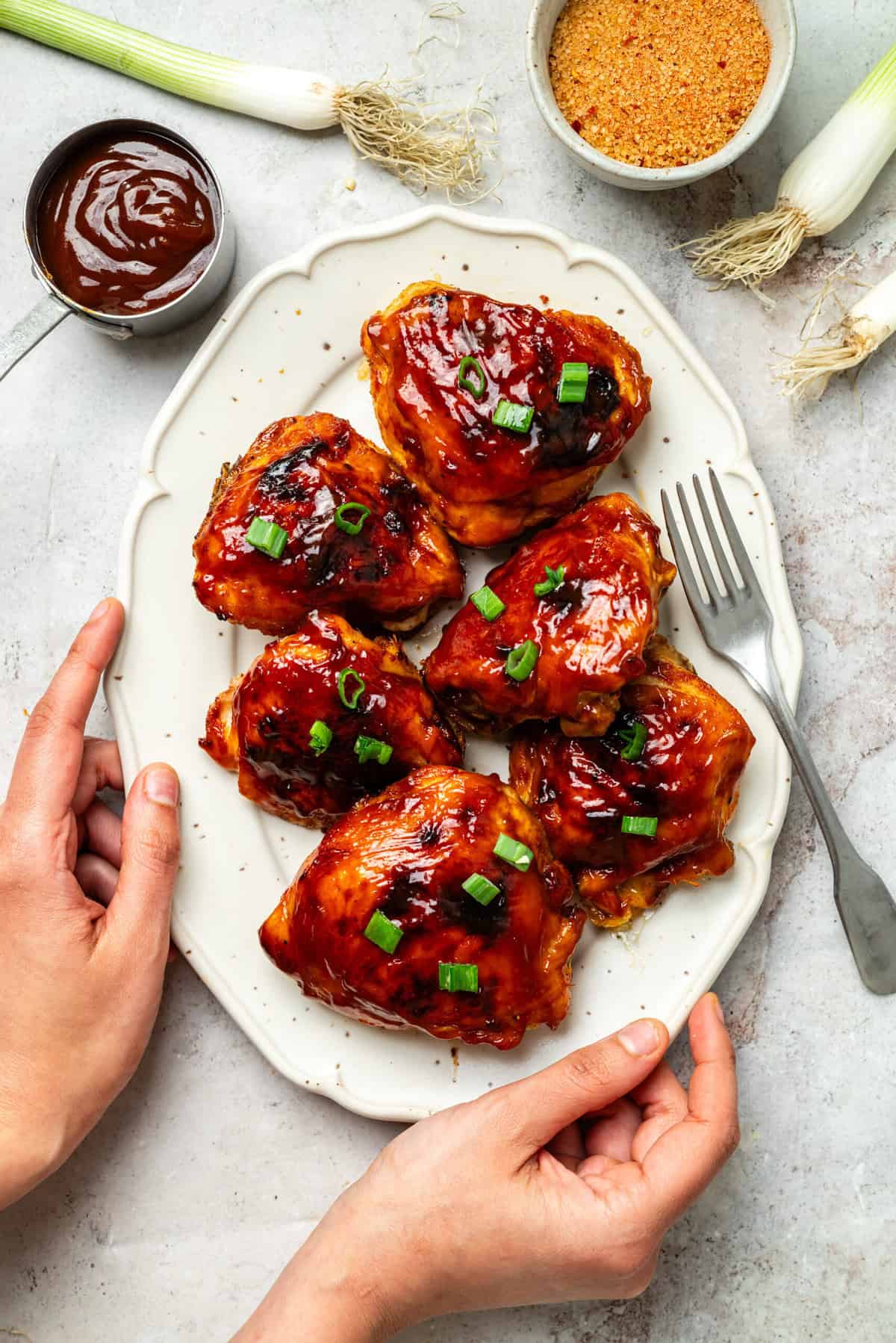 Juicy barbecue chicken drumsticks glazed with sauce, garnished with chopped green onions, served on a white plate, with ingredients and sauce nearby.
