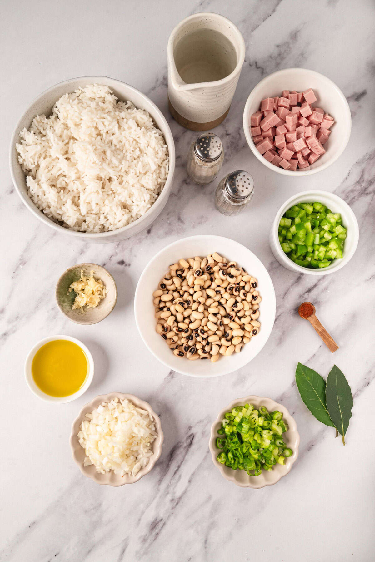 Cooked rice, black-eyed peas, diced celery, ham, chopped onions, minced garlic, olive oil, ginger, and spices, all arranged on a white marble surface for a healthy and flavorful meal preparation.